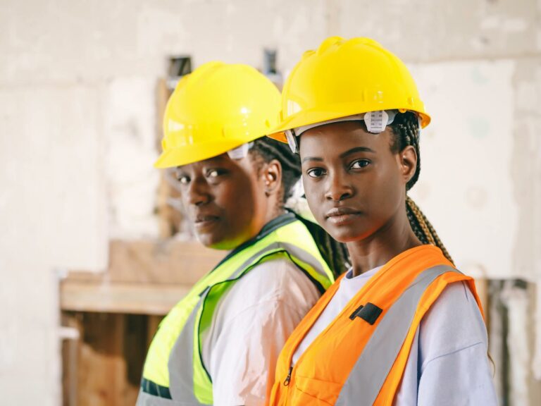 Two female construction workers wearing hardhats and vests, focusing confidently indoors.
